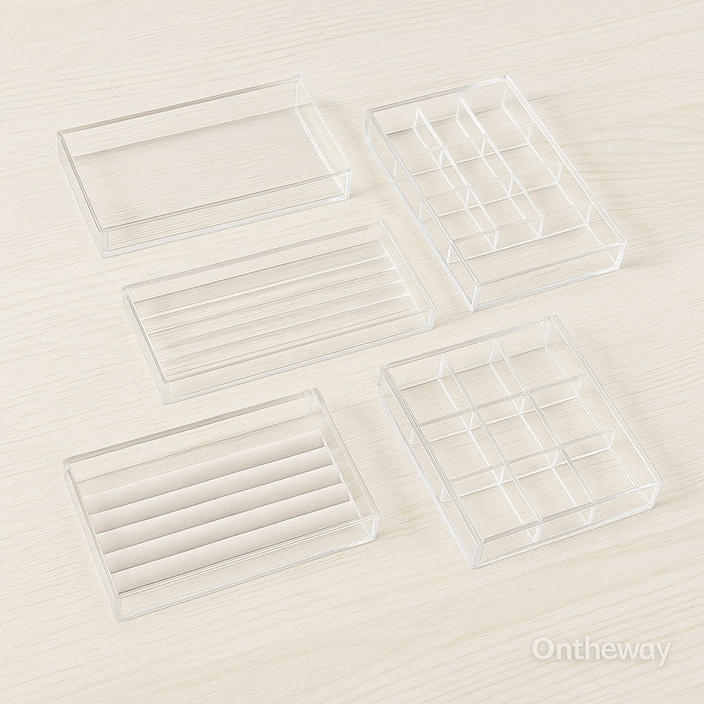 A photograph displays five clear acrylic jewelry trays in different layouts—including grid compartments, open flat trays, and section dividers—arranged neatly on a light wooden surface with a subtle Ontheway watermark. The transparent acrylic showcases a modern and minimal display style suitable for wholesale buyers.