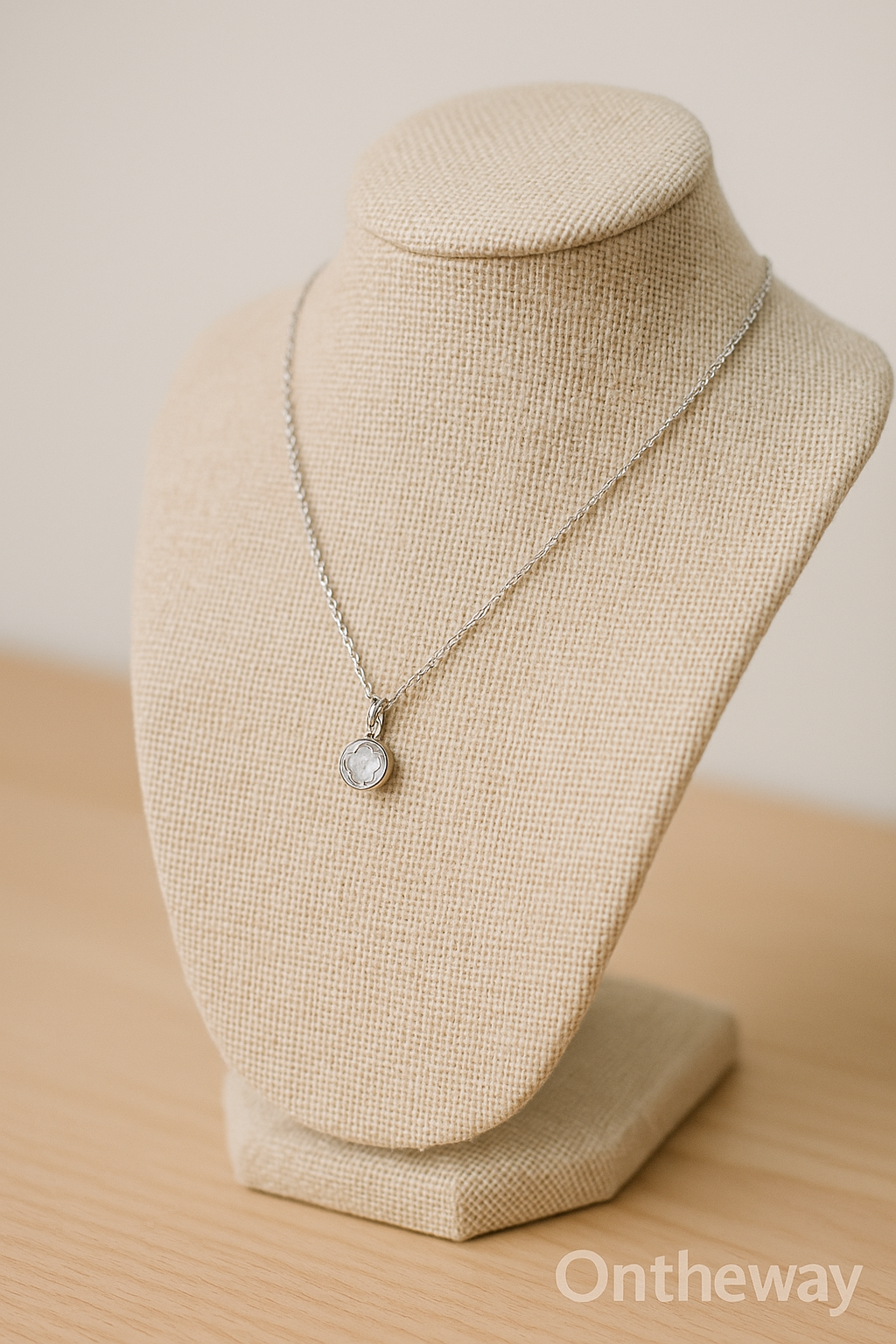 A close-up digital photograph of a beige linen jewelry display stand holding a silver necklace with a round gemstone pendant, placed on a light wooden surface under soft neutral lighting with a subtle Ontheway watermark, showcasing refined retail presentation.