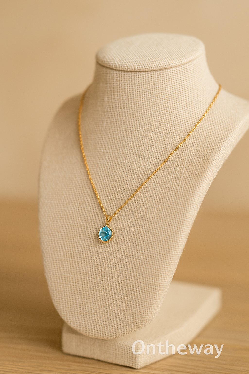 A close-up photograph of a beige linen necklace stand jewelry display showcasing a gold chain with a round blue gemstone pendant, placed on a light wooden surface under soft warm lighting with a subtle Ontheway watermark.